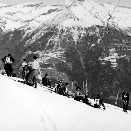 Ein Schwarz-Weiß-Foto zeigt Skifahrer und Zuschauer auf einem verschneiten Berghang. Die Teilnehmer tragen Skiausrüstung, und einige halten Skistöcke. Ein Skifahrer hat die Nummer '2' auf seiner Jacke.