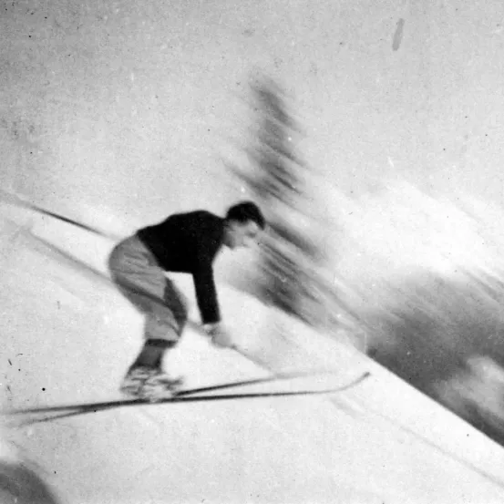 Ein Schwarz-Weiß-Foto eines Skifahrers, der einen verschneiten Hang hinabfährt und einen Skistock hält.