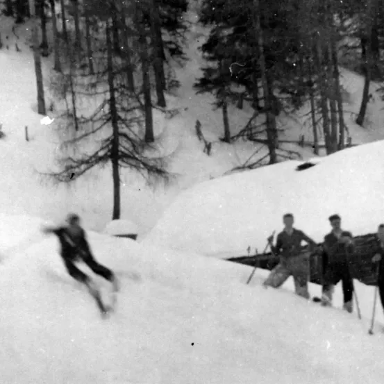 Ein Schwarz-Weiß-Foto eines Skifahrers, der einen verschneiten Hang hinunterfährt, mit drei Männern im Hintergrund, die Skistöcke halten.