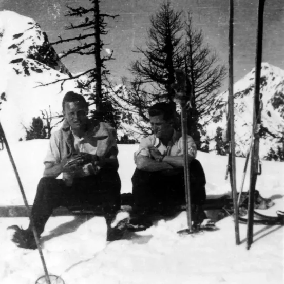 Zwei Männer sitzen im Schnee mit Skistöcken, umgeben von Bäumen und Bergen.