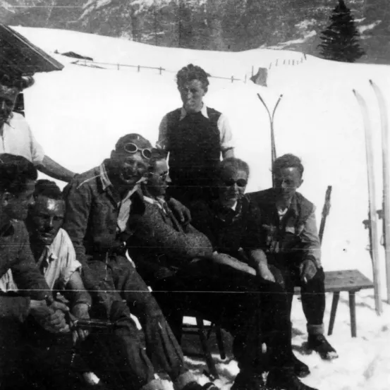 Eine Gruppe von Menschen in Winterkleidung sitzt auf einer Bank im Schnee, einige mit Skiern im Hintergrund.