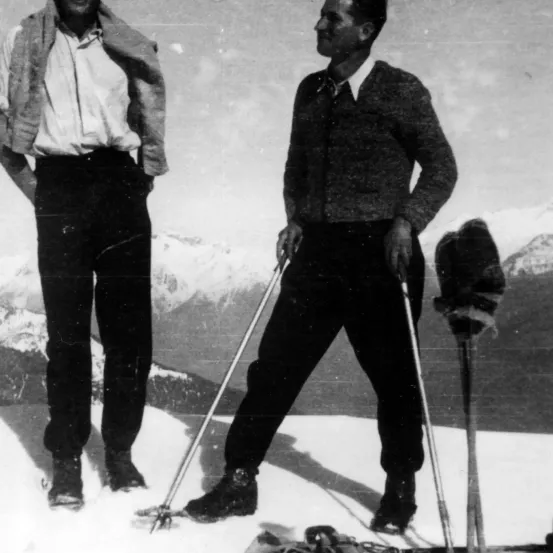 Zwei Männer in Winterkleidung stehen auf schneebedecktem Boden, halten Skistöcke in der Hand, mit Bergen im Hintergrund.