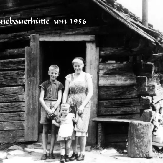 Schwarz-Weiß-Foto von etwa 1956. Eine dreiköpfige Familie steht vor einer Blockhütte. Die Frau und die beiden Kinder lächeln.