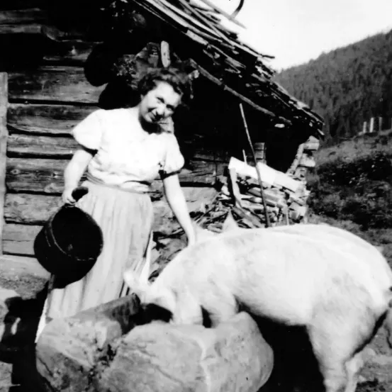 Eine Frau in einem weißen Kleid füttert ein weißes Schwein mit einem Eimer in einer ländlichen Umgebung mit einem Holzhaus im Hintergrund.