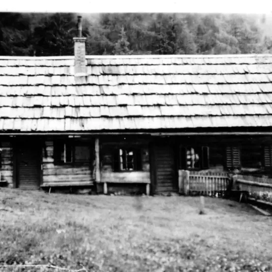 Ein Schwarz-Weiß-Bild einer Blockhütte mit Schornstein, umgeben von Bäumen und Gras.
