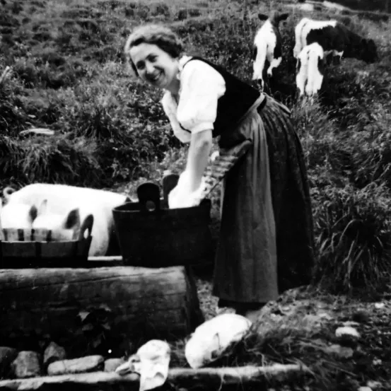 Ein Schwarz-Weiß-Bild zeigt eine Frau in einem Kleid draußen mit Nutztieren. Sie lächelt und hält einen Eimer. Im Hintergrund sind zwei Kühe zu sehen.