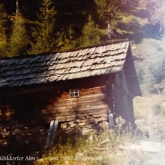 Ein altes Holzhaus namens Muhldorfer Alm, um 1980 abgebrannt, steht an einem Hang mit felsigem Gelände und üppiger Vegetation.