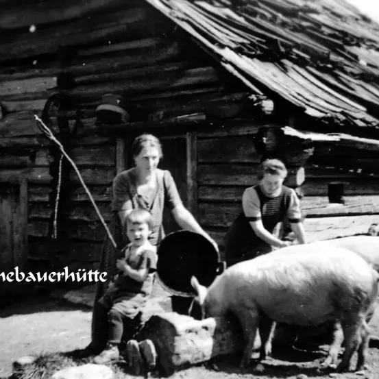 Ein Schwarz-Weiß-Bild einer Frau, eines Jungen und eines Schweins vor einer Blockhütte. Die Frau hält einen Eimer und einen Stock. Der Junge sitzt neben dem Schwein.