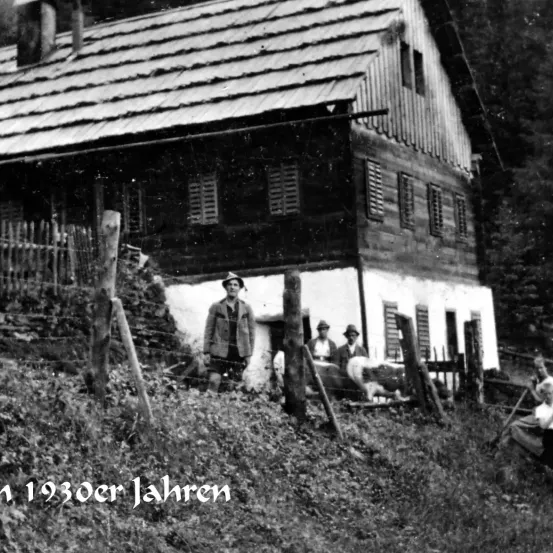 Ein Schwarz-Weiß-Foto zeigt ein Haus mit einem Mann davor. Das Haus hat ein geneigtes Dach, mehrere Fenster und einen Zaun davor. Vor dem Haus stehen mehrere Personen. Die Bildunterschrift lautet '1930er Jahren.'
