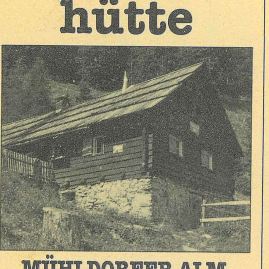 Ein monochromes Bild einer kleinen Blockhütte mit einem geneigten Dach und Schornstein. Sie steht an einem Hang mit umliegender Grünfläche. Die Hütte heißt 'Muehl Dorfer Alm'.