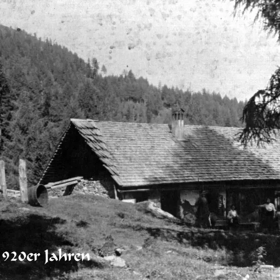 Ein Schwarz-Weiß-Foto zeigt ein altes Berghaus mit einem Strohdach und einem Schornstein. Das Haus ist von Bäumen umgeben und hat einen rustikalen Charakter. Ein Datum, 920 Jahre, ist unter dem Foto geschrieben.