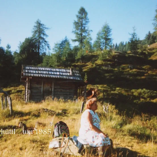 Eine Frau sitzt in einem Grasbereich, wahrscheinlich in einem Berg, um 1985. Ein Rucksack ist neben ihr platziert. Hinter ihr steht ein Holzhaus.