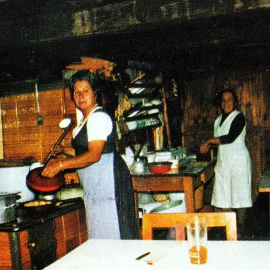 Eine alte Küche mit Holzwänden. Eine Frau kocht am Herd und hält einen Kochlöffel. Eine andere Frau steht bei einem Tisch mit einer Schüssel, beide in Schürzen. Der Raum hat Holzregale, Töpfe und einen Stuhl.