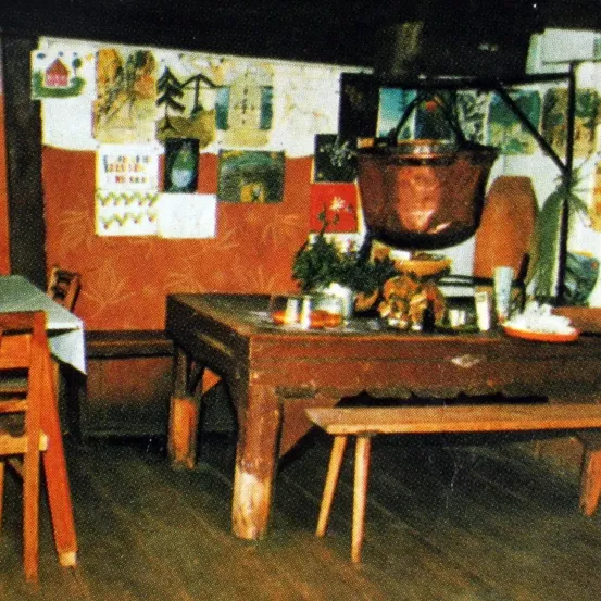 Ein Holztisch mit verschiedenen Gegenständen darauf, wie einem Topf, Tassen und einer Schüssel, steht in einem Raum. Ein Kupfertopf hängt von der Decke. Eine Bank befindet sich auf der linken Seite und ein Holzstuhl auf der rechten. Die Wand ist mit verschiedenen Zeichnungen und Gemälden geschmückt.