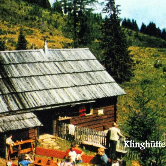 Ein Holzhaus mit rostigen Dach und Schornstein steht in einem Berggebiet. Menschen sitzen um einen Tisch vor dem Haus, während ein Mann in der Nähe steht.