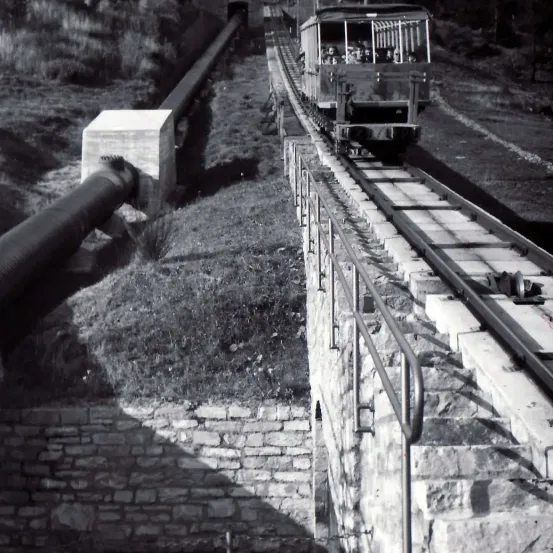 Ein Schwarz-Weiß-Bild zeigt einen Zug auf einer Strecke mit Fahrgästen. Der Zug ist auf einer Ziegelstruktur erhöht. Eine große Rohrleitung befindet sich links am Boden.