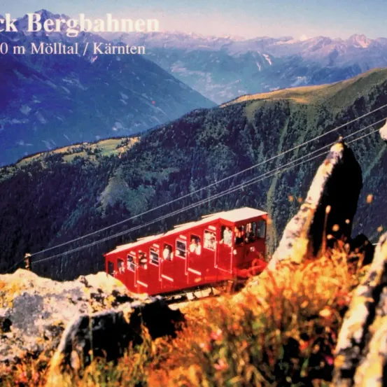 Eine rote Seilbahn an einem steilen Berghang in Molltal, Karnten, Österreich. Die Seilbahn ist an einem Kabel befestigt und von grüner Vegetation umgeben.