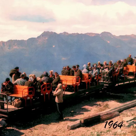 Ein Vintage-Foto eines Zuges mit Passagieren, die auf orangefarbenen Stühlen auf einer Bahnstrecke sitzen, mit Bergen im Hintergrund. Das Jahr 1964 ist handgeschrieben in der unteren rechten Ecke.