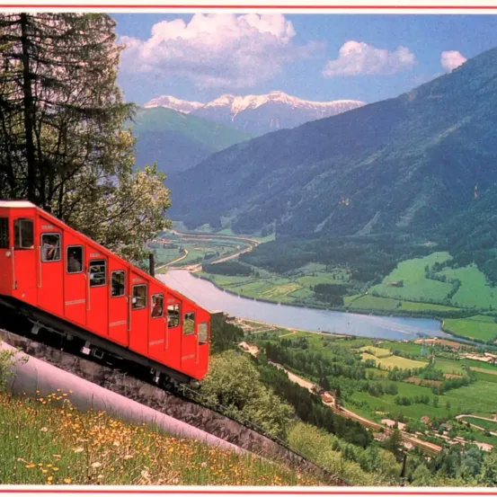 Ein roter Zug fährt einen steilen Berghang hinauf, mit einem üppigen grünen Tal und einem Fluss darunter. Berge und Bäume im Hintergrund.