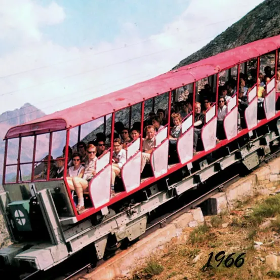 Ein Vintage-Bild von 1966 zeigt eine rote Seilbahn mit vielen Passagieren im Inneren, die einen Berg hinauf fährt, mit einer malerischen Kulisse im Hintergrund.