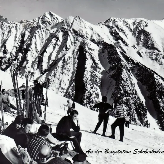 Schwarz-Weiß-Foto eines verschneiten Berges mit sitzenden und stehenden Personen. Einige haben Skier an und andere haben Skistöcke. In der Ferne sind Berge sichtbar. Die Bildunterschrift lautet 'An der Bergstation Schoberboden'.