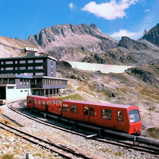 Ein roter Zug fährt auf einer Bergstrecke mit einem großen Gebäude im Hintergrund. Das Gebäude hat viele Fenster und befindet sich in der Nähe eines Berges mit einer Steinmauer.