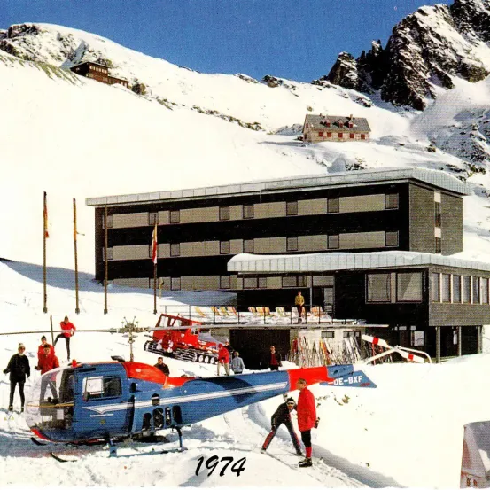 Vintage-Foto von 1974, das eine verschneite Landschaft mit einem Hotel, einem Hubschrauber und Menschen zeigt. Das Gebäude hat ein geneigtes Dach und Flaggen. Im Vordergrund stehen Menschen in der Nähe eines Hubschraubers.