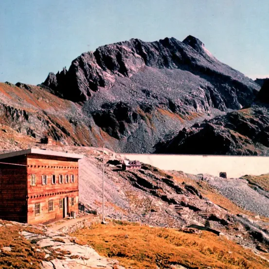 Ein Holzgebäude mit mehreren Fenstern steht vor einer bergigen Kulisse. Die Landschaft ist rau mit sichtbarem felsigem Gelände. In der Ferne ist ein See mit einem Damm zu sehen.