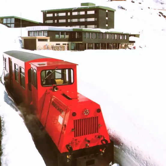 Ein roter Zug mit grünem Dach fährt durch tiefen Schnee vor einem grünen Gebäude mit vielen Fenstern.