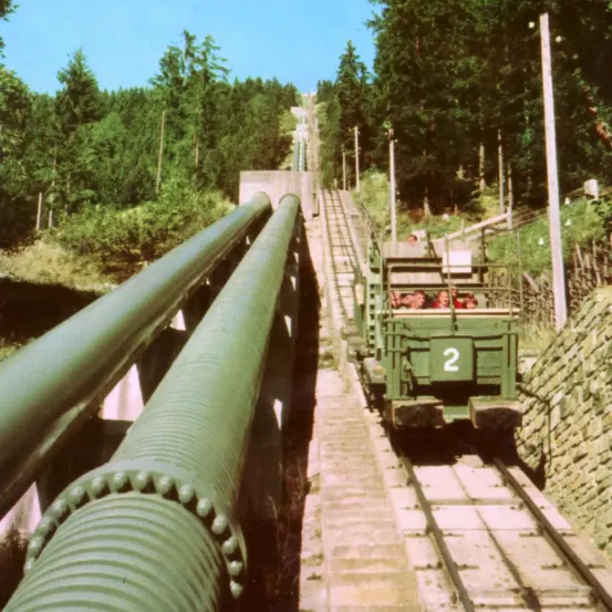 Ein grüner Zug mit der Nummer 2 fährt auf einer Bahnstrecke durch einen Wald, begleitet von zwei großen grünen Rohren.