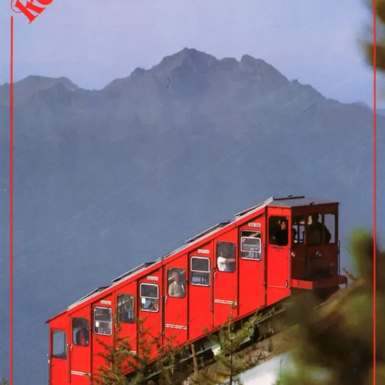 Eine rote Seilbahn fährt bergauf, mit Passagieren, die durch die Fenster zu sehen sind. Im Hintergrund befinden sich Berge unter einem klaren Himmel.