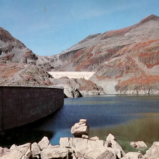 Ein Foto eines Damms mit Bergen und Felsen. Der Damm ist grau und die Berge sind braun.