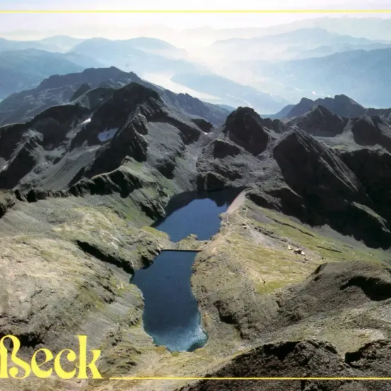 Luftaufnahme einer Bergregion mit zwei Seen in der Mitte. Die Berge sind felsig und mit spärlicher Vegetation bedeckt. Der Himmel ist klar mit einigen Wolken.