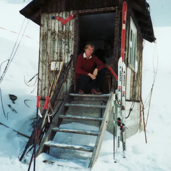 Ein Mann sitzt in einem kleinen hölzernen Kabineneingang mit Skiern, die an den Wänden lehnen, er ist in einem roten Pullover und Hose gekleidet.