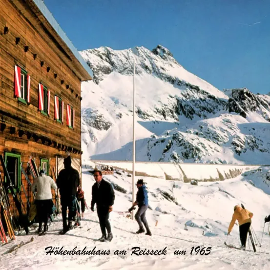 Eine Gruppe von Menschen steht vor einem Gebäude mit Skiern, die dagegengelehnt sind. Im Hintergrund befinden sich schneebedeckte Berge. Das Bild ist mit 'Hohenbahnhaus am Reisseck um 1965' beschriftet.