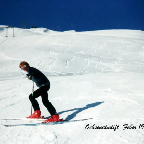 Ein Skifahrer in roten Skischuhen fährt im Februar einen schneebedeckten Hang hinunter. Das Bild ist mit 'Ochsenalmlift' und dem Jahr 19 beschriftet.