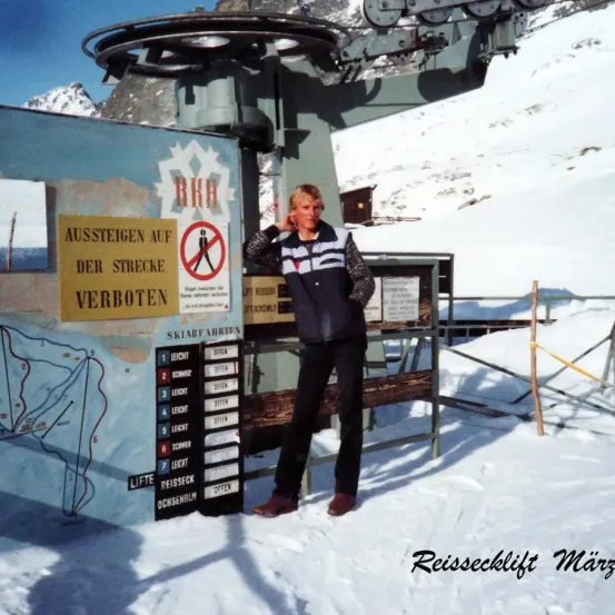 Ein Mann steht neben einer Skiliftstation. Das Schild hinter ihm besagt 'Aussteigen auf der Strecke verboten' und 'Lift reisen'. Eine Karte mit nummerierten Wegen ist an der Wand montiert.