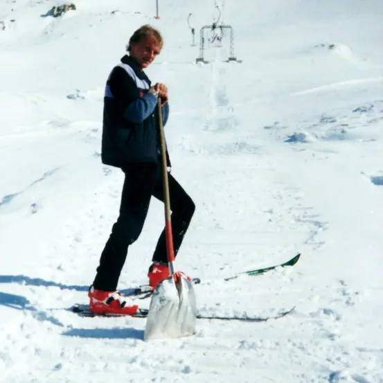 Ein Mann steht auf Skiern, hält eine Schaufel und räumt Schnee in einem verschneiten Bereich.