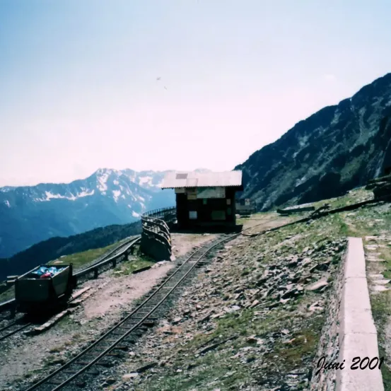 Ein Bergbahnhaltepunkt mit einem kleinen Gebäude auf einem steilen Hang, umgeben von felsigem Gelände und Bergen unter einem klaren Himmel.