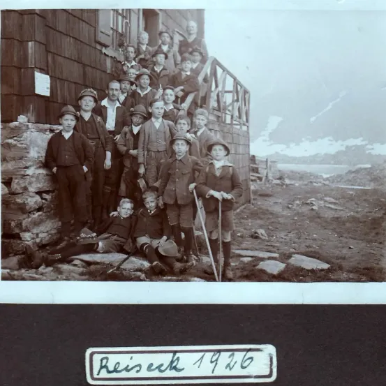 Eine alte Schwarz-Weiß-Foto von einer Gruppe von Menschen vor einem Holzgebäude mit einem Berg im Hintergrund. Das Jahr 1926 steht auf einem Etikett am unteren Rand.