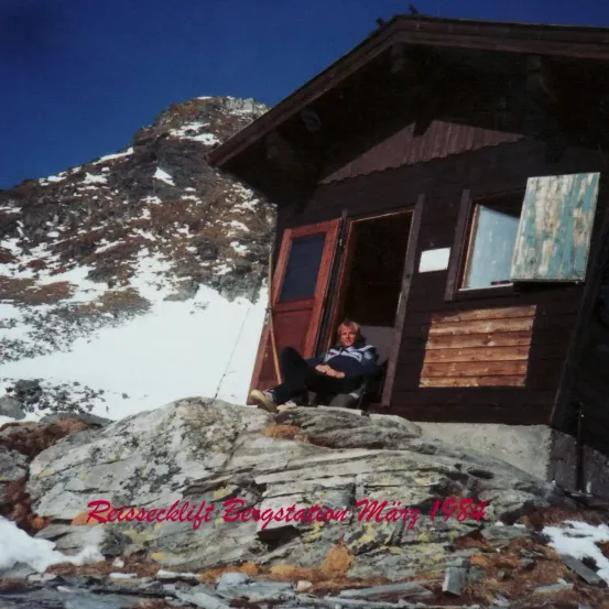 Eine Frau sitzt in der Tür einer Holzhütte mit schneebedeckten Bergen im Hintergrund. Darunter steht der Text 'Reissecklift Bergstation März 1984'.