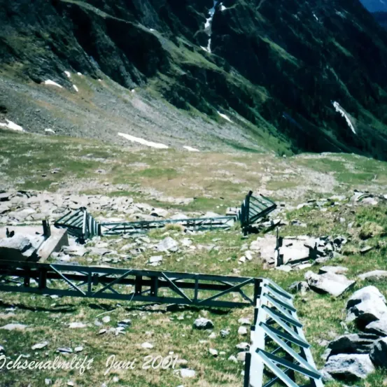 Berglandschaft mit grünem Gras und Felsen, einem zerbrochenen Zaun und einem Wasserzeichen auf Deutsch, das 'Schsenadlerlift Juni 2001' lautet.