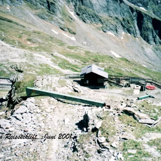 Eine Berglandschaft mit einer kleinen Hütte mit einem geneigten Dach, einer grünen Rohrleitung am Boden und einem Bergpfad, der zu ihr hinaufführt. Die Szene ist vom Juni 2001 datiert.