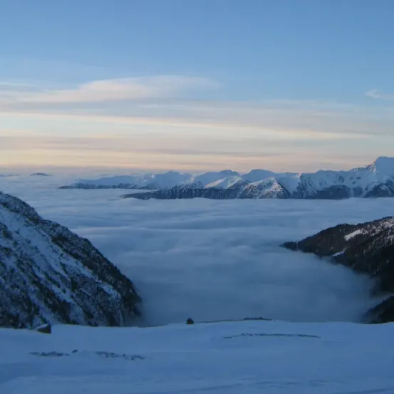 Ein schneebedeckter Berggipfel mit einem Meer aus Wolken darunter, umgeben von anderen Bergen, unter einem blauen Himmel mit Wolken.