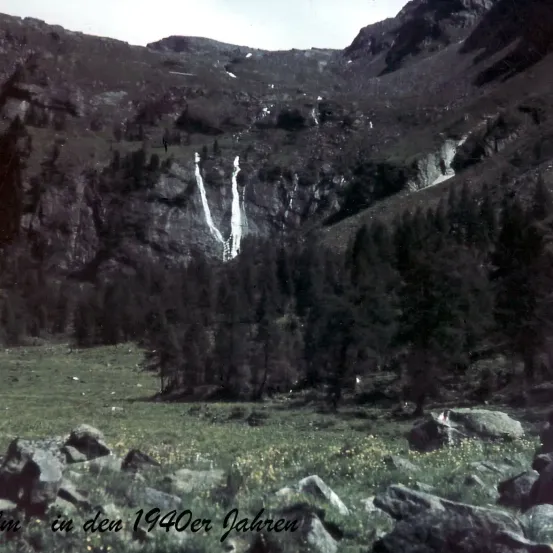 Eine Berglandschaft aus den 1940er Jahren mit einem Wasserfall, der über die Felsen strömt, umgeben von dichtem Wald und felsigem Gelände.