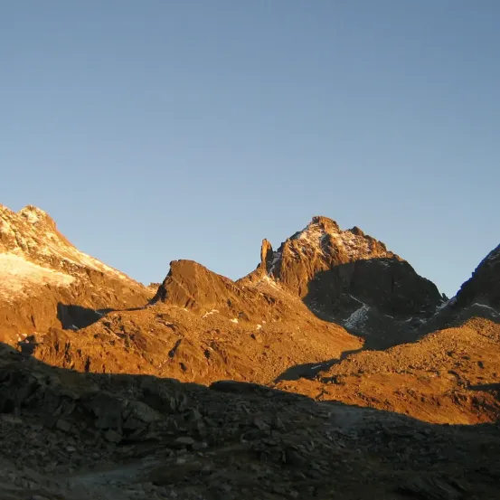 Eine malerische Ansicht eines Gebirgszugs mit einem goldenen Schimmer, der von der untergehenden Sonne geworfen wird. Schneebedeckte Gipfel sind sichtbar, und das raue Gelände ist rau.