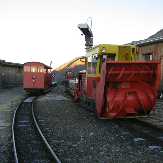 Zwei Züge auf einer Schiene, einer ist ein kleiner roter Zug mit einem gelben Dach und der andere ist ein großer roter und gelber Zug mit einem Schneepflug.