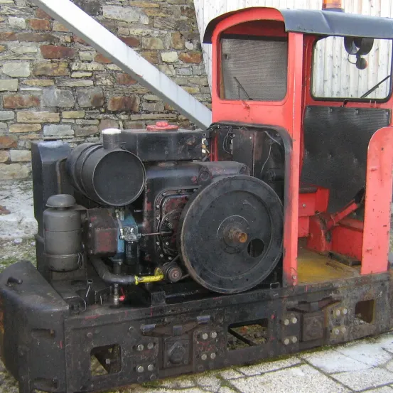 Eine alte rote Lokomotive steht vor einer Ziegelmauer. Sie hat einen schwarzen Korper und rote Verkleidung. Der Motor hat ein großes Rad und einen schwarzen Auspuff.