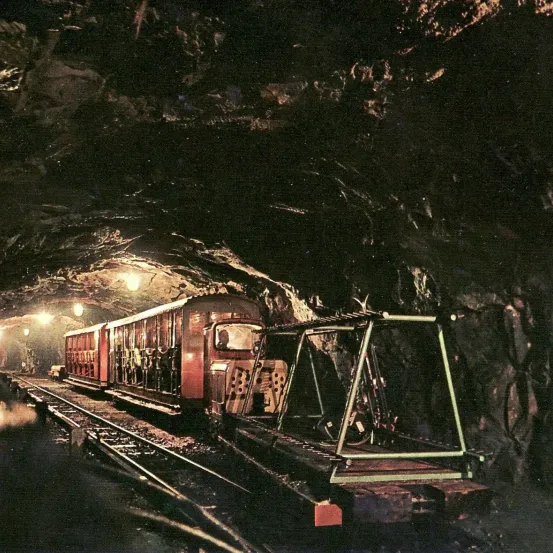 Ein alter Zug befindet sich in einem dunklen Tunnel, der von einigen Lichtern beleuchtet wird. Der Zug hat viele Fenster, und eine Person ist drinnen. Die Tunnelwände sind felsartig.