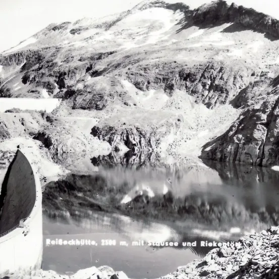 Ein Schwarz-Weiß-Bild eines Berges mit einem See im Vordergrund, der Text lautet 'Reisbockhütte, 2300 m, mit Stausee und Riekentor.'